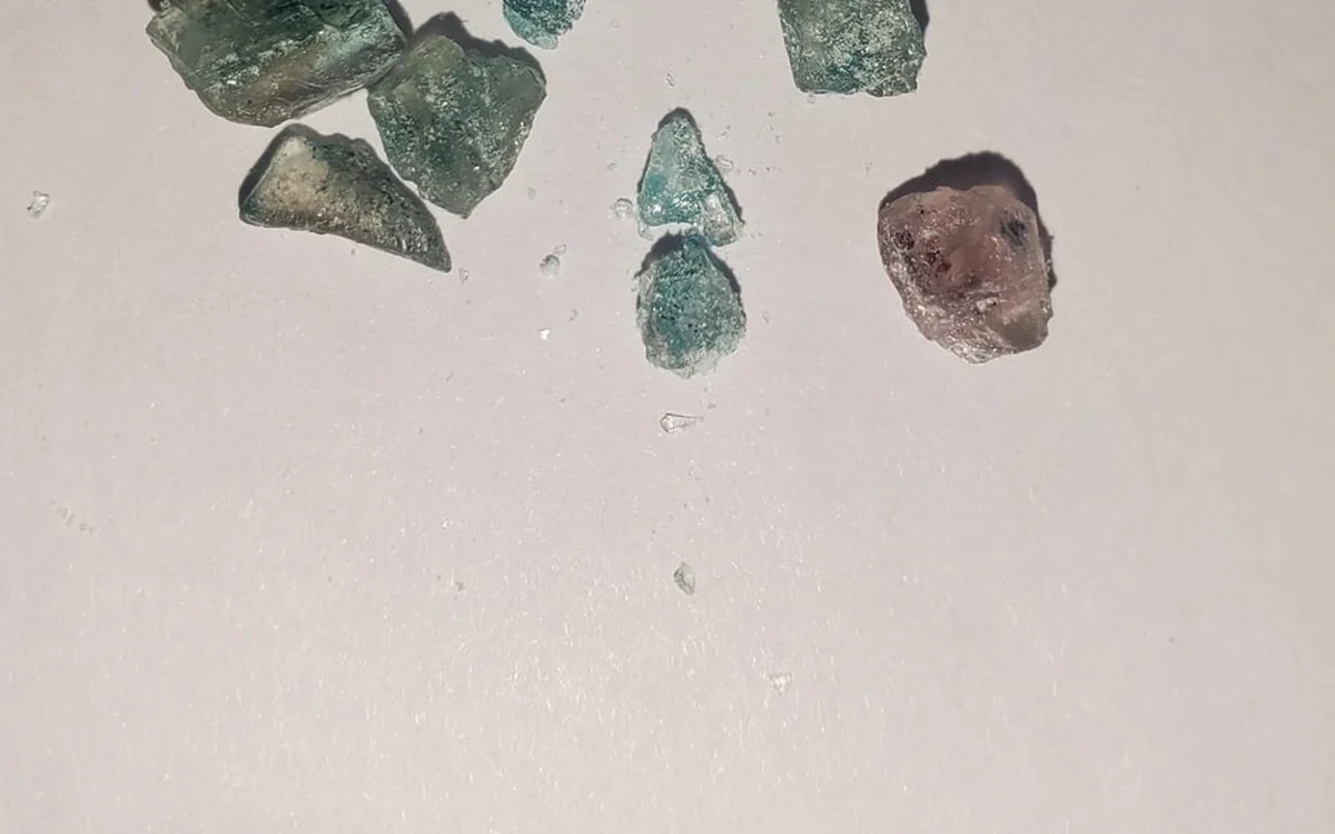 Six MDMA crystal shards on a white surface, several blue green and one brown purple
