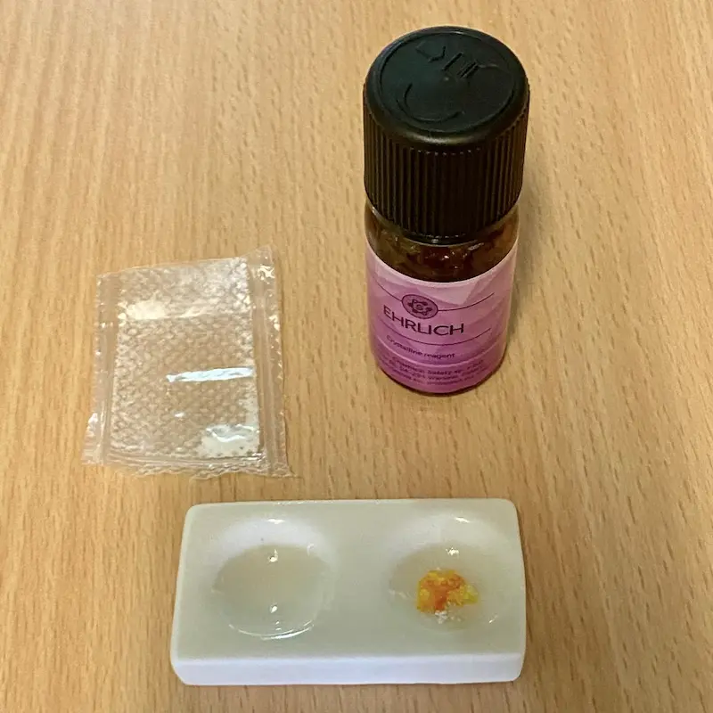 A bottle of Ehrlich reagent beside a small bag of powder and a white spot plate with two wells: one stays pale, the other turns bright orange