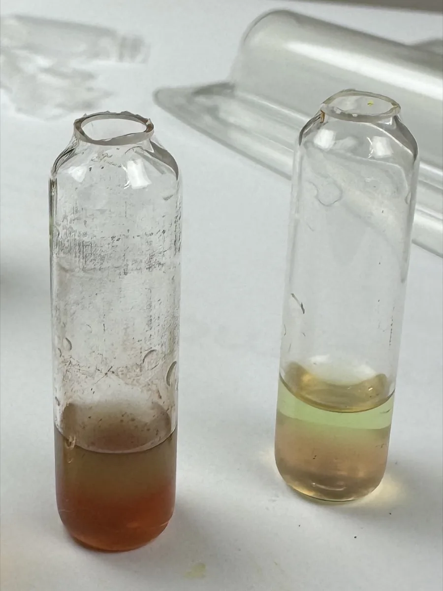 Two opened one-liquid cocaine quantity ampoules side by side, one orange-brown and one pale yellow with a thin green layer
